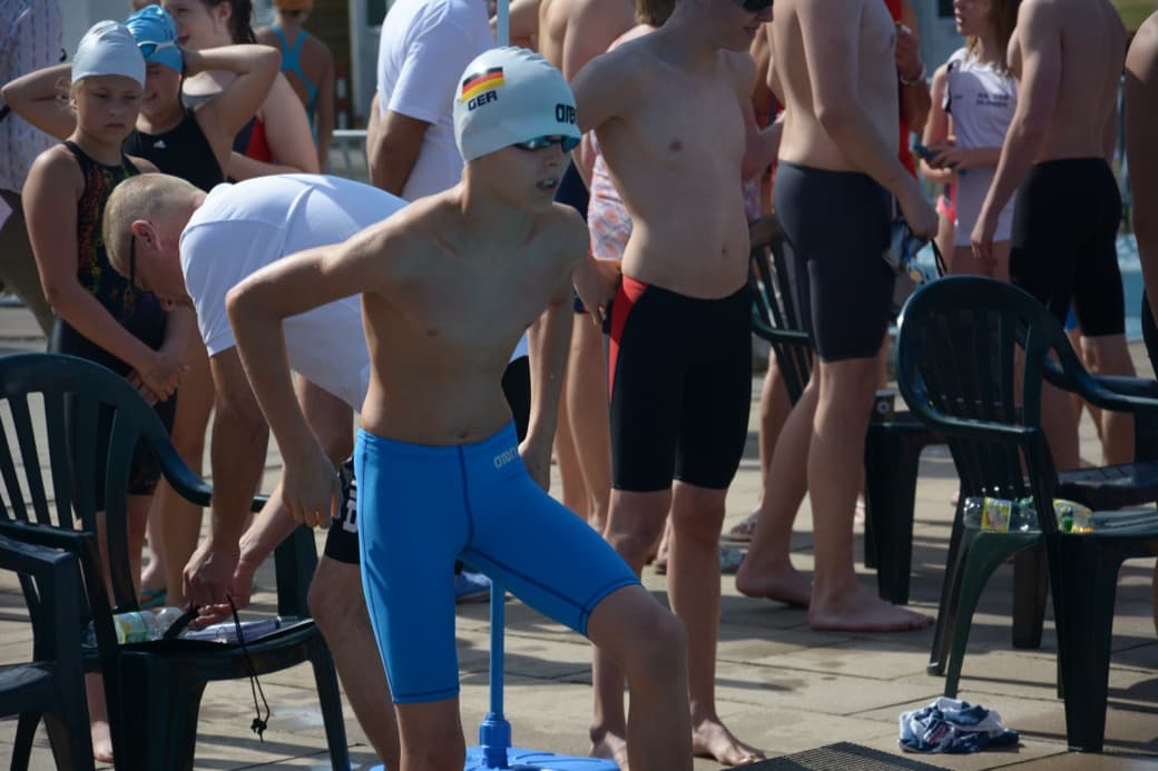 A young swimmer in a white GER cap and blue jammers standing poolside among other competitors at an outdoor meet.